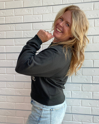 A woman with blonde hair smiling and posing in front of a white brick wall, wearing a black long-sleeve cropped sweatshirt and a light blue denim mini skirt.