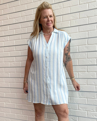 Woman wearing a light blue and white striped dress standing against a white brick wall.