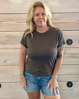 Woman wearing a brown t-shirt and denim shorts standing against a wooden wall.