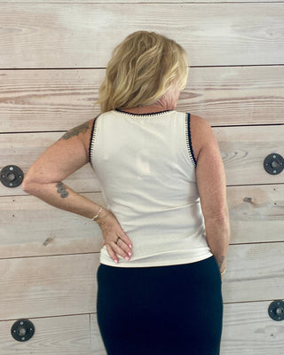 Person wearing a white sleeveless top and dark pants against a wooden wall.