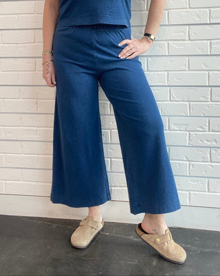 Woman wearing a blue sleeveless top and wide-leg pants against a white brick wall.