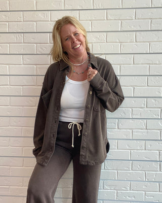 A woman posing in front of a white brick wall wearing a dark brown button-up shirt jacket over a white tank top, dark brown wide-leg pants, and leopard print shoes.