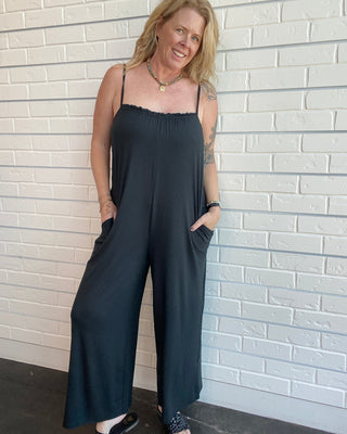 Woman wearing a black jumpsuit standing against a light brick wall.