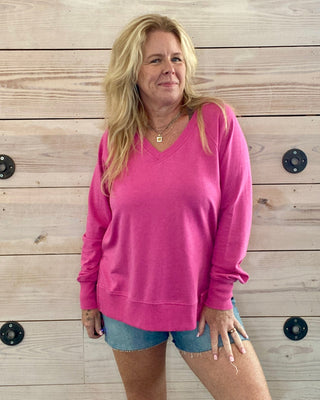 Woman wearing a pink sweater and denim shorts standing against a wooden wall.