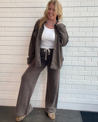 A woman stands against a white brick wall wearing a matching taupe brown lounge set consisting of a button-up cardigan and wide-leg pants, styled with a white tank top, black drawstring pants, and leopard print shoes.