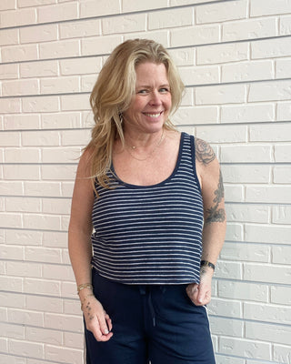 Woman wearing a striped tank top and dark pants standing against a light brick wall.