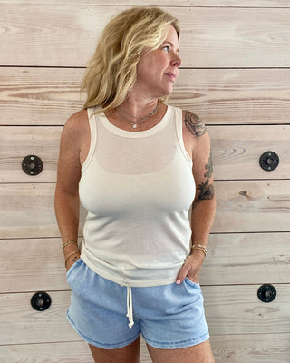 Woman wearing a cream tank top and light blue shorts standing against a wooden wall.