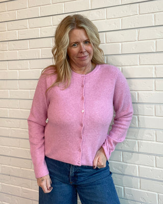 Woman wearing a pink cardigan and blue jeans standing against a light brick wall.