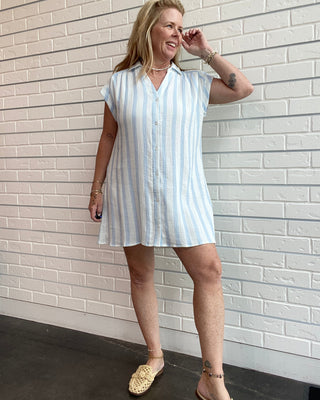 Woman wearing a light blue and white striped dress against a white brick wall.