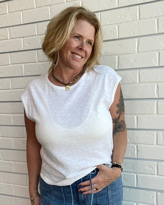Woman wearing a white top and blue jeans standing against a light brick wall.