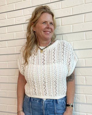 Woman wearing a white crochet top and blue jeans standing against a beige brick wall.