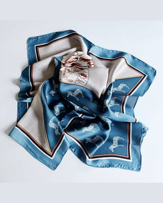 A square silk scarf with a blue and beige design, featuring white zebra prints and a central illustration of a woman, laid flat on a light gray background.