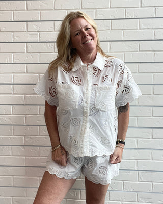 Woman wearing a white embroidered shirt and shorts against a white brick wall.