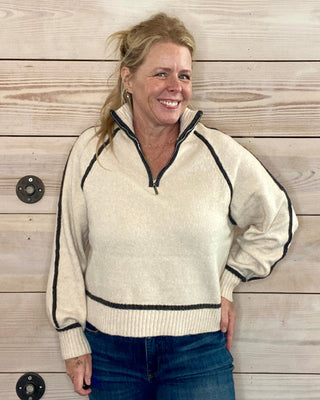 Woman wearing a cream-colored sweater with black trim against a wooden wall.