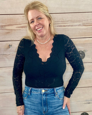Woman wearing a black long-sleeve top and blue jeans standing against a wooden wall.