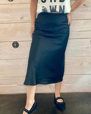 Person wearing a white t-shirt with text and a black skirt against a wooden background
