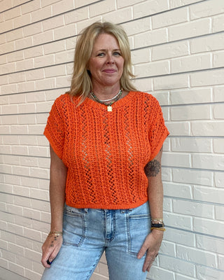 Woman wearing an orange knitted top and blue jeans standing against a light brick wall.