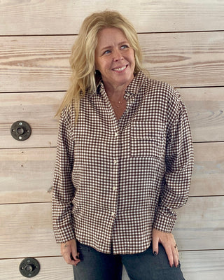 Woman wearing a plaid shirt and jeans standing against a wooden wall.