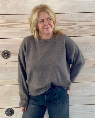 Person wearing a gray sweater and dark jeans standing against a wooden wall.
