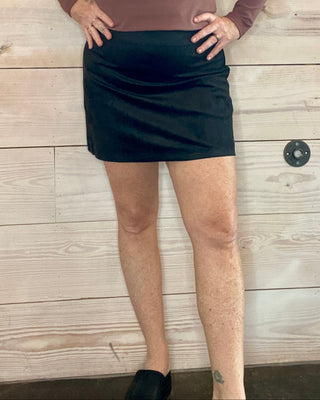 Woman wearing a brown long-sleeve top and black skirt against a wooden wall.