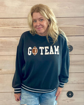 Person wearing a black sweatshirt with 'GO TEAM' text against a wooden wall.