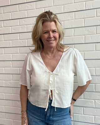 Woman wearing a white blouse and blue jeans standing against a light brick wall.