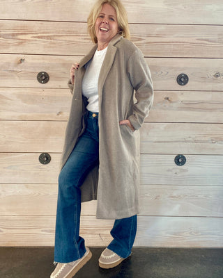 Person wearing a long beige coat over a white shirt and blue jeans, standing against a wooden wall.