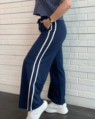 Woman wearing a striped top and navy pants with white stripes against a light brick wall.