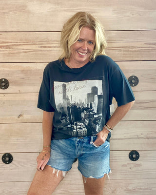 Woman wearing a grey graphic t-shirt and denim shorts standing against a wooden wall.