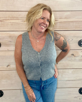 Woman wearing a gray vest and blue jeans standing against a wooden wall.