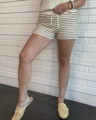 Woman in a sleeveless top and shorts standing against a white brick wall.