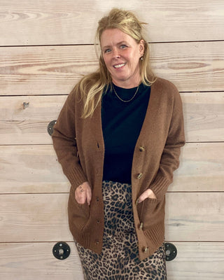 Woman wearing a brown cardigan, black top, and leopard print skirt against a wooden wall.