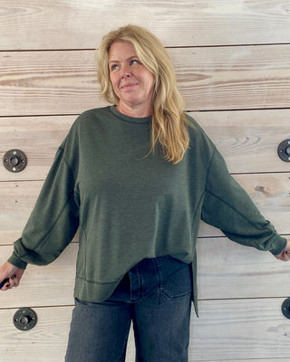 Person wearing a green sweatshirt and dark jeans standing against a wooden wall.