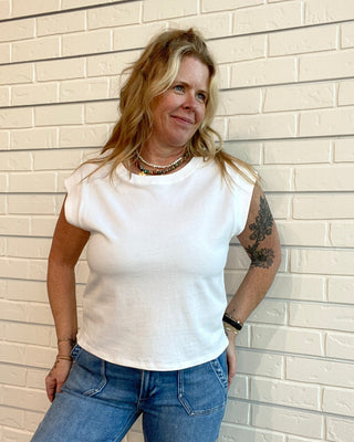 Woman wearing a white t-shirt and blue jeans standing against a light brick wall.