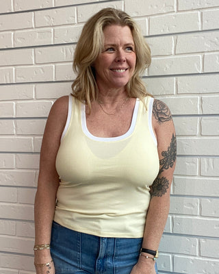 Woman wearing a yellow tank top and blue jeans standing against a light brick wall.
